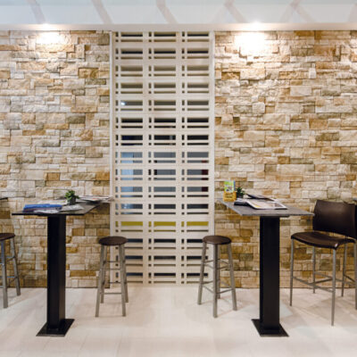 Espacio de trabajo con revestimiento de pared efecto piedra natural en tonos beige y ocre, mostrando piezas irregulares superpuestas que crean una textura 3D. Mesas altas negras con taburetes metálicos y celosías decorativas blancas complementan el acabado rústico del gres porcelánico.