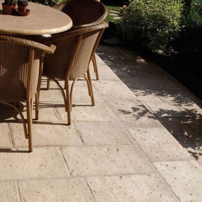 Terraza exterior pavimentada con baldosas de gres porcelánico antideslizante en tonos ámbar cálidos, rodeada de vegetación y plantas en macetas, con sombras de follaje proyectadas sobre el suelo.