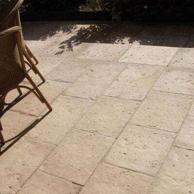 Terraza exterior pavimentada con baldosas de hormigón en tonos beige y gris, textura rústica imitación piedra natural, con sillas de ratán y sombra de vegetación al fondo.
