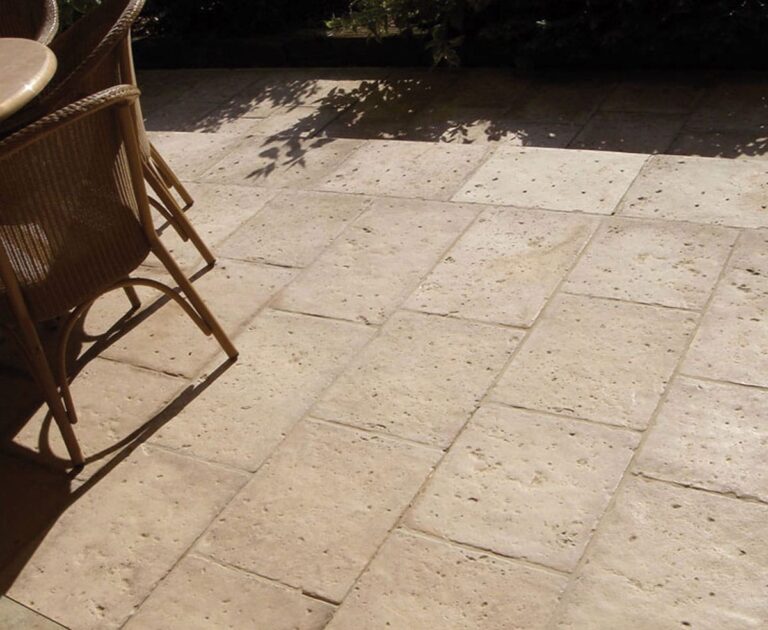 Terraza exterior pavimentada con baldosas de hormigón en tonos beige y gris, textura rústica imitación piedra natural, con sillas de ratán y sombra de vegetación al fondo.