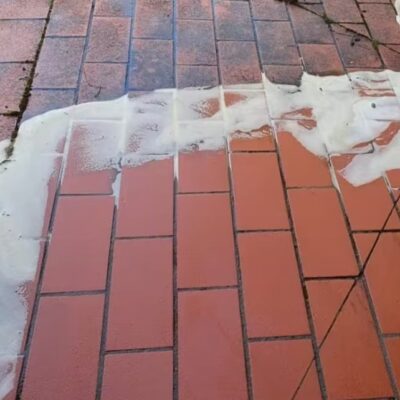 Pavimento exterior de suelo cerámico en tonos rojizos y naranjas con patrón de colocación a tresbolillo. La mitad superior muestra el material limpio y la inferior con manchas blancas de suciedad, demostrando el contraste de limpieza. Formato rectangular con juntas grises visibles.