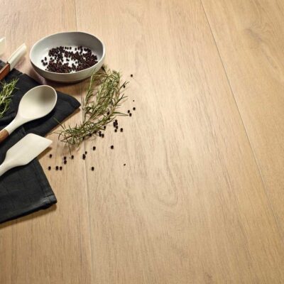 Conjunto de herramientas culinarias con mango de madera, hierbas aromáticas frescas y semillas de mostaza negra dispuestas sobre superficie de madera clara. Incluye cucharas, espátulas y un cuenco cerámico con especias.