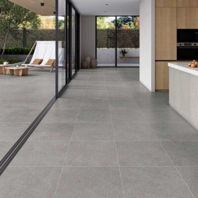 Cocina y zona de estar con suelo porcelánico efecto piedra en tonos grises, amplios ventanales que abren a terraza exterior equipada con tumbonas y zona de estar. El gres porcelánico continúa tanto en interior como en el espacio exterior, creando una transición visual fluida entre ambas áreas.