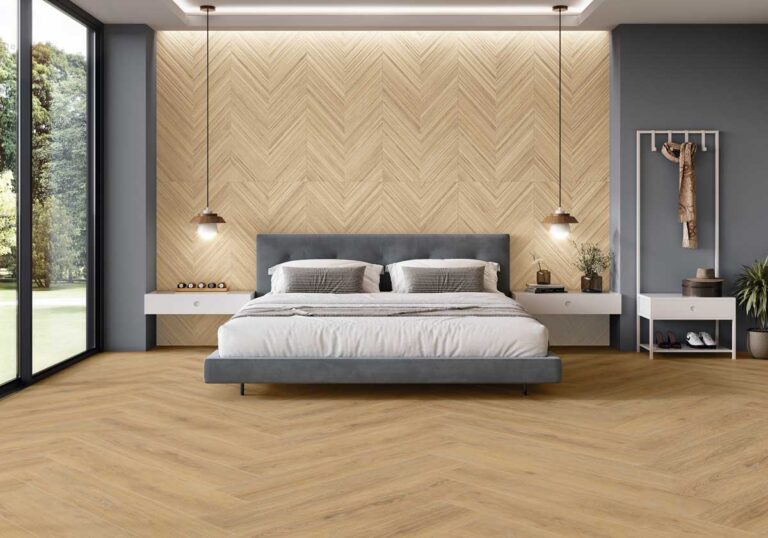 Dormitorio contemporáneo con gres porcelánico imitación madera en tonos miel claro, instalado en patrón de espiga. Pared de fondo con revestimiento cerámico texturizado en tonos naturales, cama tapizada gris oscuro con mesillas flotantes blancas, iluminación colgante industrial y acceso a jardín mediante cristalera.