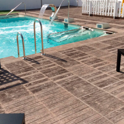 Terraza de piscina exterior con suelo de gres porcelánico en acabado madera clara, junto a una piscina turquesa con cascada y zona de spa jacuzzi, rodeada de mobiliario y valla blanca de seguridad.