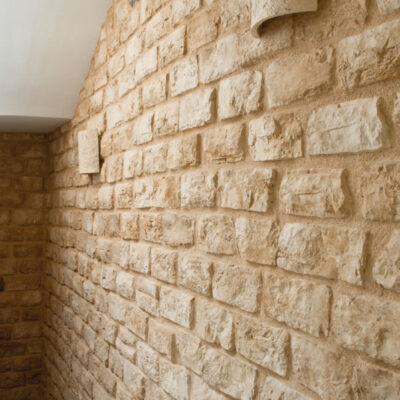 Pasillo interior con revestimiento de pared Piamonte Verniprens en tonos beige claro, que imita textura de ladrillo con acabado rústico. Suelo con patrón geométrico chevron blanco y gris visible en primer plano.
